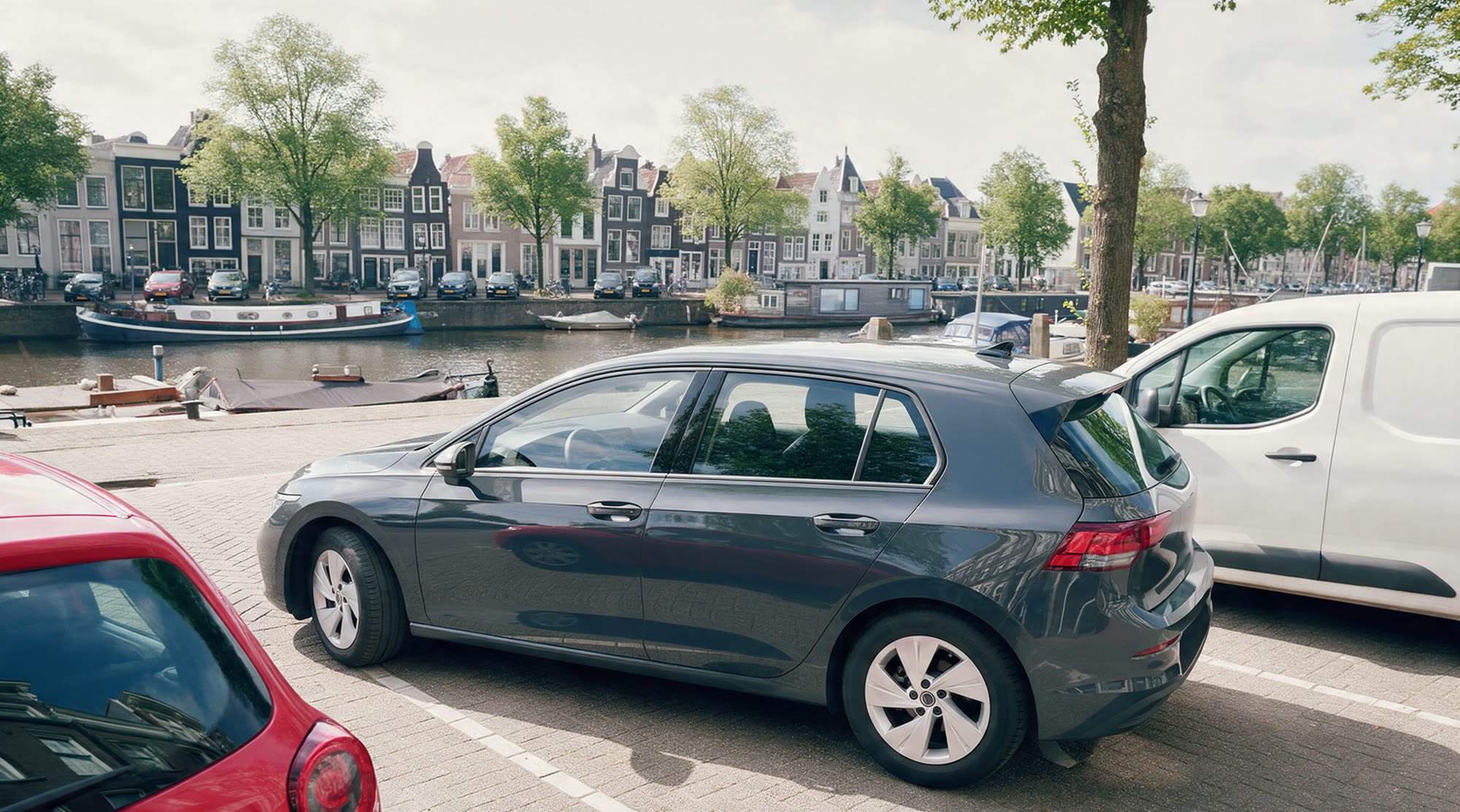 De lege lesauto staat halverwege het inparkeren in een ruime parkeerplek tussen een rode auto en een witte bestelbus aan een gracht